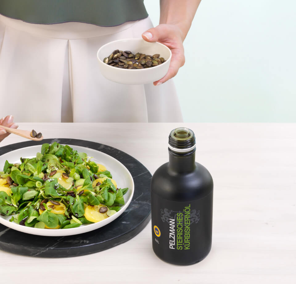 Eine Person streut Kürbiskerne aus einer Schüssel auf einen Kartoffelsalat mit original steirischem Pelzmann Kürbiskernöl. Daneben steht eine Flasche Pelzmann Steirisches Kürbiskernöl.